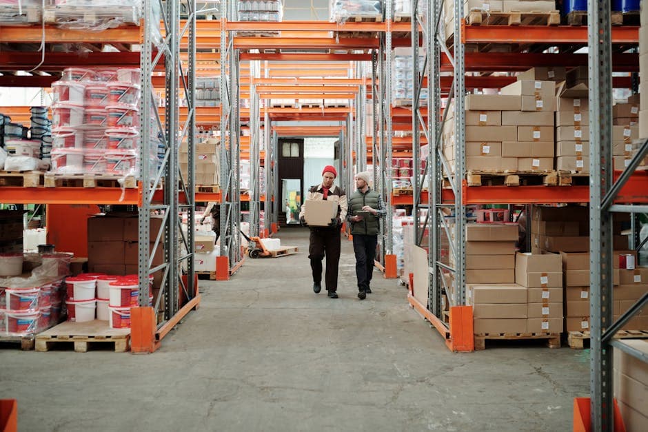 Workers walking along aisle in warehouse surrounded by shelves stocked with boxes and products
